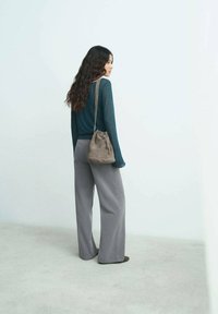 Pantalones de pierna ancha grises, una blusa de manga larga en tono teal y un pequeño bolso tipo bucket de ante marrón. La modelo está de pie contra una pared clara, con el cabello ondulado.