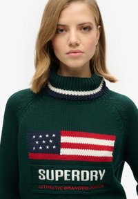 Pull en tricot vert avec un col montant, ornée d'un motif de drapeau américain et du texte "SUPERDRY" en accents blancs et rouges.