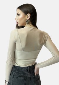 Transparente Langarmbluse in Beige mit taillierter Silhouette, runder Halslinie und nahtlosem Design. Kombiniert mit dunklen Denim-Shorts.