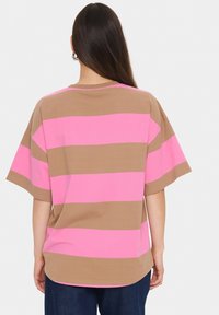 Femme avec de longs cheveux portant une chemise ample à manches courtes avec de larges rayures horizontales roses et beiges, se tenant de dos.