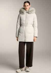 Palton puffer de culoare gri deschis, cu glugă căptușită cu blană, talie fixată cu brău și cusături orizontale. Modelul poartă pantaloni negri și adidași.