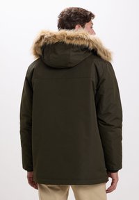 Uomo di spalle che indossa un parka verde scuro con cappuccio foderato in pelliccia e pantaloni beige, su uno sfondo bianco semplice.