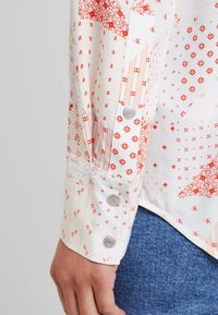 Gros plan sur une manche de chemise blanche avec des motifs géométriques et floraux rouges, des boutons-pression en argent et une main reposant près d'un jean en denim bleu.