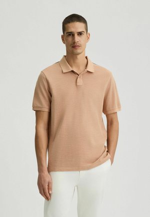 Hellbeige Polo-Shirt aus strukturiertem Stoff. Mit klassischem Kragen, kurzen Ärmeln und zwei Knöpfen zu schließen. Getragen mit weißen Hosen.