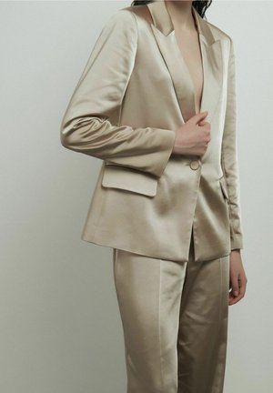 Personne portant un costume en satin beige brillant avec un blazer à un bouton et un pantalon droit assorti, posant avec une main sur le blazer.