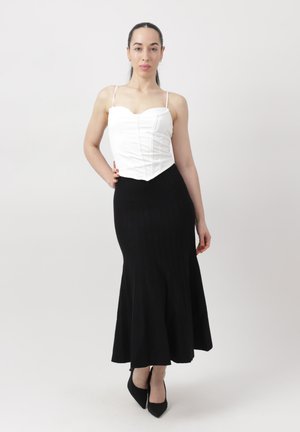 Kobieta stoi z ręką na biodrze, ubrana w biały bezrękawnik typu gorset, czarną długą spódnicę i czarne szpilki na jednolitym tle.