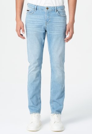 Jean en denim bleu clair avec coupe droite, cinq poches, décoloration subtile et une patch en cuir marron au dos de la taille. Associé à des baskets blanches.