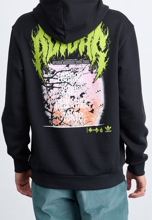 Person trägt schwarzen Hoodie mit neongrünem grafischem Text und pink-orangener abstrakter Kunst auf dem Rücken, die Städte für die Frühling-Sommer-Tourdaten 2025 auflistet.