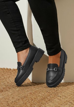 Mocasines de cuero negro con una suela gruesa y texturizada, que cuentan con una correa frontal con detalles de herrajes plateados y un diseño de punta redondeada.