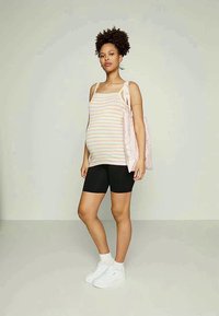 Mulher grávida em pé, vestindo uma t-shirt de listras, calções pretos, ténis brancos e a segurar uma mala de pano rosa, em frente a um fundo simples.