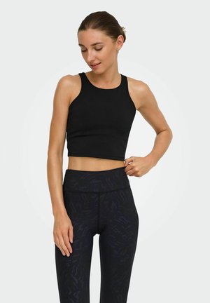 Kvinde iført sort ærmeløs cropped top og mønstrede højtbemandede leggings, står og justerer taljen med en lys baggrund.