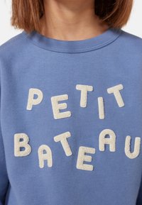 Modrá mikina s texturovanými krémovými písmeny, které hlásají "PETIT BATEAU." Má kulatý výstřih a měkkou tkaninu. Detailní pohled.
