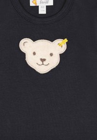 T-shirt in cotone blu navy con applicazione di un volto di orso in feltro beige, ricamo nero e un nastro giallo all'accufronte destro.
