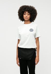 Biela bavlnená tričko s krátkymi rukávmi, s okrúhlym grafickým logom v tmavomodrej farbe. Nosené s čiernymi nohavicami a zodpovedajúcim čiernym pouzdrom.