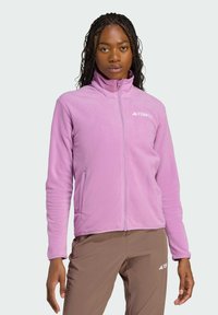Helles lila Fleece-Jacke mit durchgehendem Reißverschluss, hohem Kragen und seitlichen Taschen. Ist mit einem kleinen weißen Logo auf der Brust ausgestattet.