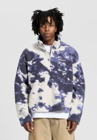 Fleece pullover in cream and navy blue, featuring a quarter-zip neckline and textured, cloud-like pattern throughout. Logo on chest.