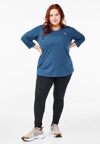 Blue, lightweight athletic top with three-quarter sleeves and a round neckline, paired with black leggings and beige sneakers.