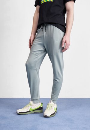 Dolna część ciała osoby ubranej w jasnoszare joggery Nike i białe sneakersy z neonowo zielonym logo Nike, stojącej na niebieskiej podłodze przy białej ścianie.