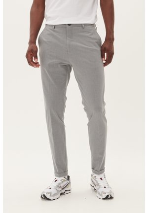 Homme portant un pantalon gris clair slim et des chaussures de sport blanches, debout devant un fond uni.