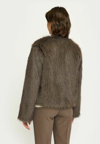 Jachetă din blană sintetică de culoare maro, cu o formă pătrată și mâneci lungi, având o textură moale, asemănătoare cu părul lung și un tiv scurt.