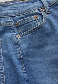 Blauwe spijkerbroek van denim met een klassieke pasvorm, stevig materiaal, koperen klinknagels en gestikte accenten langs de zakken en naden.