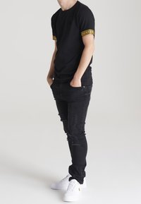 Camiseta negra con la marca "SikSilk" en amarillo en las mangas, combinada con unos vaqueros negros desgastados y zapatillas blancas. Outfit casual, corte moderno.