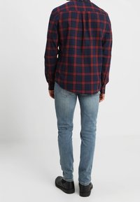 Marineblauw en rood geruit flanellen overhemd met een afgeronde zoom, met knoopmanchetten, gecombineerd met lichtblauwe jeans en zwarte schoenen. Achteraanzicht.