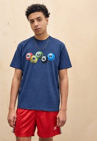 Navy blue cotton t-shirt featuring multicolored billiard balls with "CRUSH" text. Paired with red shorts featuring geometric patterns.