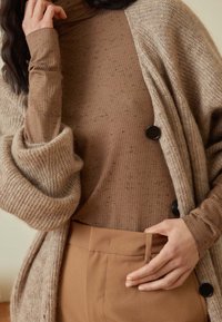 Tanstickad cardigan med överdimensionerade ärmar, knappar och en strukturerad yta, lagd över en brun ribbad turtleneck och matchande byxor.