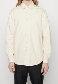 Camicia a maniche lunghe beige chiaro con colletto button-down, tasca frontale e orlo curvo. Realizzata in un tessuto morbido con dettagli di cucitura visibili.