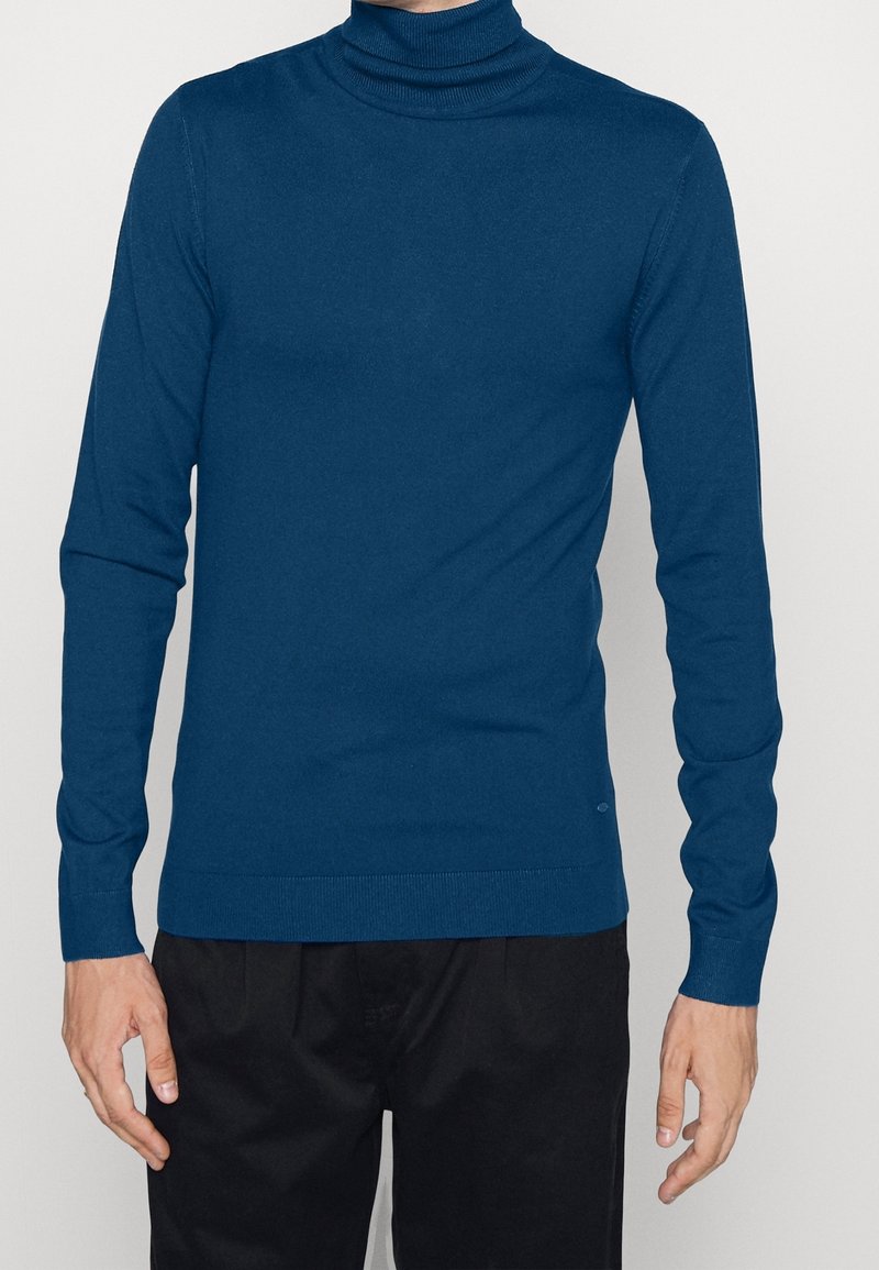 Homme portant un pull à col roulé bleu ajusté et un pantalon noir, debout avec les bras détendus le long du corps sur un fond uni.