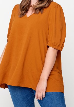 Mujer vestida con una blusa texturizada suelta de color naranja quemado, con mangas abullonadas hasta el codo, y jeans azules, de pie frente a un fondo blanco.