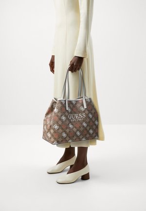 Personne en robe crème et chaussures blanches tenant un grand sac cabas Guess marron avec un logo à motif et des anses blanches.