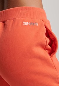 Orange mjukisbyxor i mjukt tyg, med ribbad midja och sidficka. 'SUPERDRY'-logotypen är broderad i vitt.