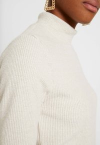 Cremfarbener Rippstrickpullover mit einem figurbetonten Design, das eine strukturierte Oberfläche und einen hohen Kragen aufweist. Betont durch eine sichtbare Schulternaht.