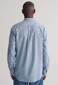 Camisa de manga larga azul clara con puños abotonados, detalle de pliegue en la espalda, dobladillo curvado y un cuello clásico en una suave tela texturizada.