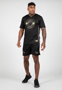Juodas sportinis marškinėlis ir šortai su auksiniais logotipo akcentais. V-kaklo dizainas, trumpomis rankovėmis ir lygia tekstūra. Dėvimi su juodais sportbačiais.
