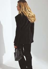 Giacca in corduroy nero con una vestibilità strutturata, abbinata a pantaloni coordinati; una borsa in pelle nera è tenuta in mano.