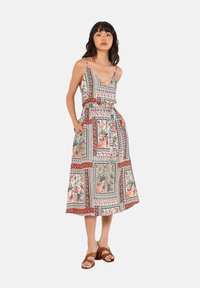 Maxikleid aus leichtem Stoff mit floralem und geometrischem Muster in Rot und Grün. Mit Spaghettiträgern und Seitentaschen. Braune Sandalen.