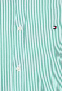 Grün-weiß gestreiftes Hemd mit Knopfverschluss, glatter Stofftextur und einem kleinen Logo mit roten, weißen und blauen Akzenten.