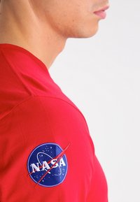 Joven con camiseta roja con el logo de NASA en la parte superior de la manga, mirando hacia la derecha sobre un fondo claro y liso.
