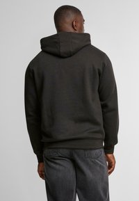 Felpa nera con cappuccio dal finish testurizzato, polsini a costine e tasca a marsupio. Indossata con jeans grigio scuro, che mostrano una vestibilità rilassata.