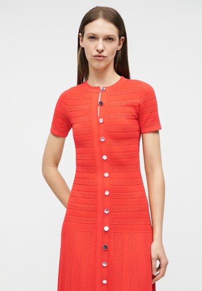 Γυναίκα με ίσια καστανά μαλλιά που φοράει ένα κόκκινο, κοντομάνικο φόρεμα με κουμπιά και υφασμένους οριζόντιους και κάθετους σχηματισμούς.
