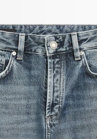 Vooraanzicht van de tailleband van een blauwe spijkerbroek met knoop, riemlussen, ritssluiting en voorzakken.