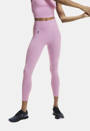 Személy nyújtja a karjait, rózsaszín sportleggingset és haspólót viselve, fekete futócipőben, egyszínű háttér előtt.