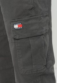 Mørkgrå cargo bukser har en sidelomme med et brodert "TOMMY JEANS" merke i blått, rødt og hvitt. Laget av slitesterkt stoff, med detaljerte sømmer synlige.