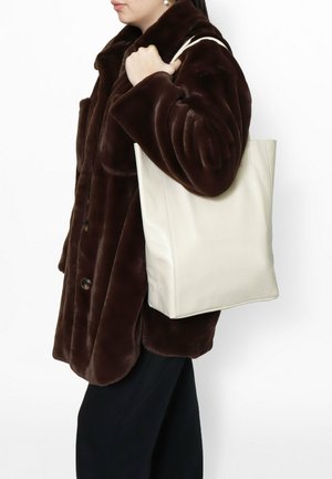 Sac fourre-tout en cuir blanc avec une forme structurée, une texture lisse et sans matériel visible. Associé à un manteau en fausse fourrure marron foncé.