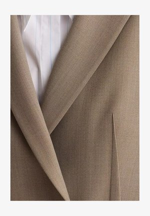 Beżowa marynarka o gładkiej fakturze, z kołnierzem szalowym i subtelnie zdobioną kieszenią. Nałożona na białą koszulę w jasnoniebieskie paski.