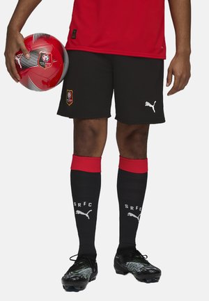 Rød atletisk skjorte med logo, sorte shorts med logo, sorte sokker med røde accenter, sorte fodboldstøvler med grønne mønstre, holder en rød og sort fodbold.