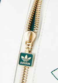 Bílý zip s kovovými zuby a táhlem zlaté barvy. Na něm je štítek s logem Adidas v zelené barvě se zlatými akcenty. Viditelné detaily texturovaného povrchu.